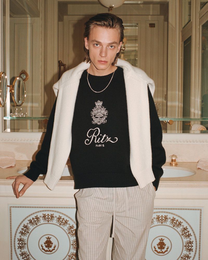 Portrait of a young adult male fashion model indoors, wearing a black crewneck sweater with the Ritz Paris logo, posing by a marble countertop.
