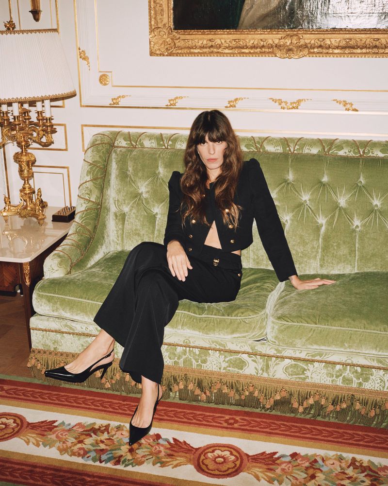 A woman in a black blazer and pants sits on a green velvet couch in an ornate room with a patterned rug and painting.