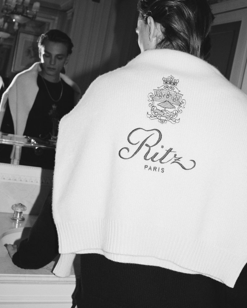 Person in mirror wearing Ritz Paris branded sweater. Indoors, the mirrored reflection reveals a second person. Black and white photography.