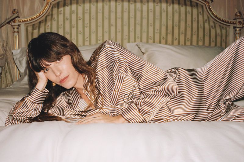 Brunette woman in striped pajamas reclining on a bed with white linens and a gold-trimmed headboard in an indoor setting.