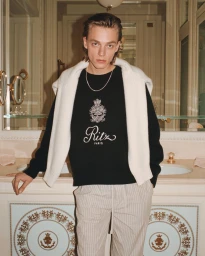 Model Michael Zielinski wearing a black Ritz Paris sweatshirt and striped trousers leaning against a marble bathroom sink.