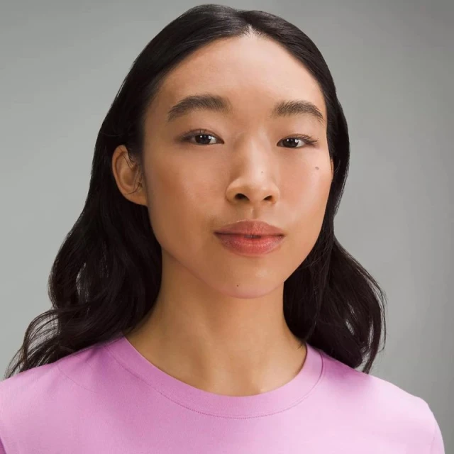Portrait of an Asian woman with dark hair and a pink t-shirt. Studio shot with a neutral grey background.