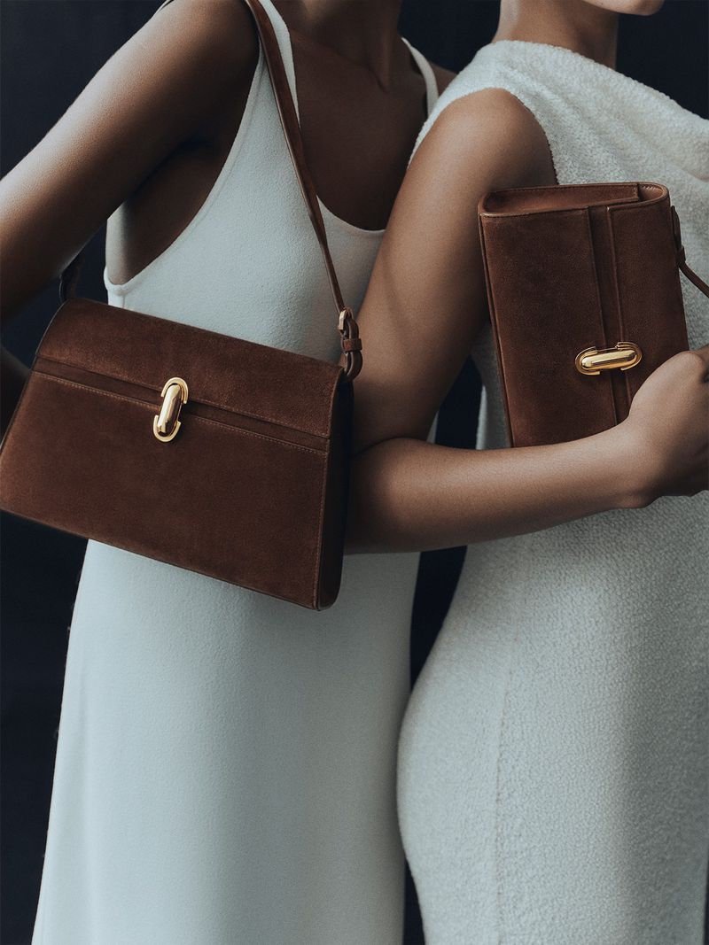 Close-up of two women wearing white dresses, each holding a brown leather handbag with gold clasp against a dark background.