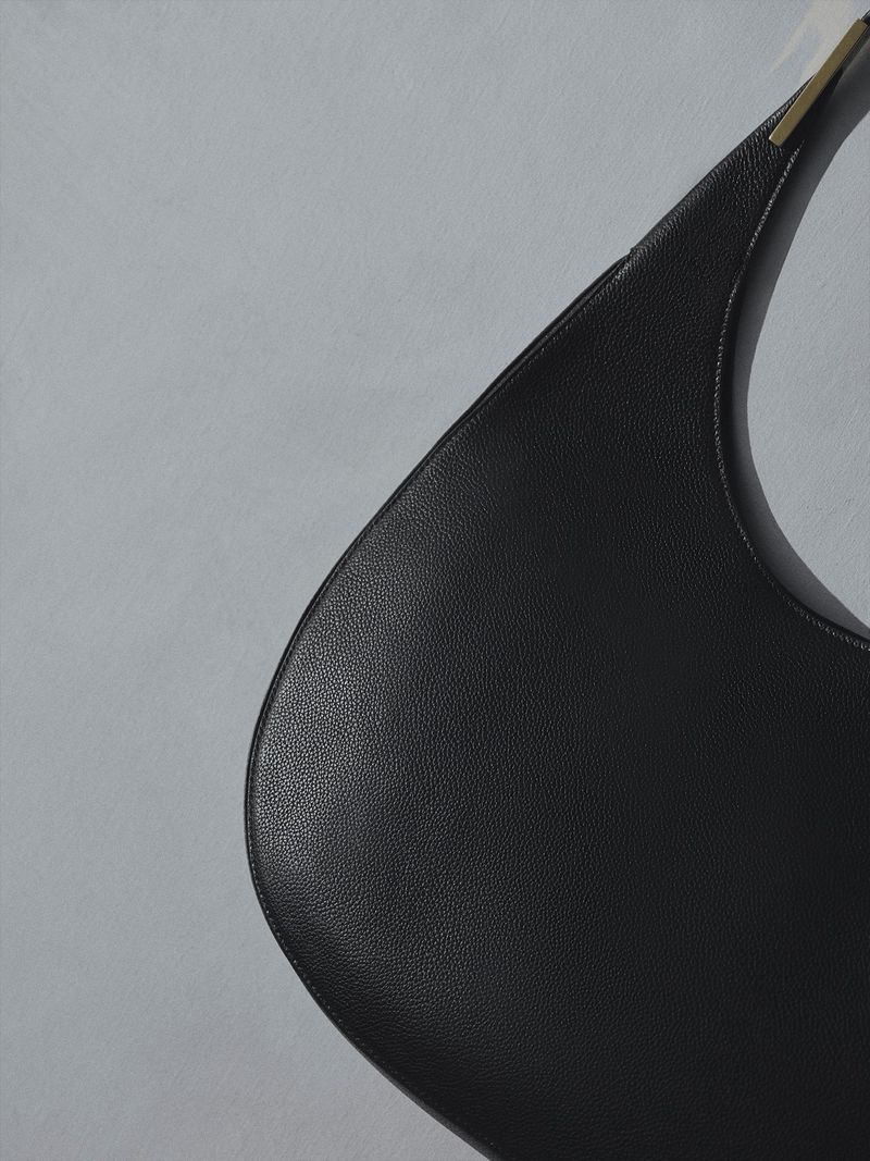 Close-up of a minimalist, black leather shoulder bag with a curved shape and a gold metal detail against a pale grey background.