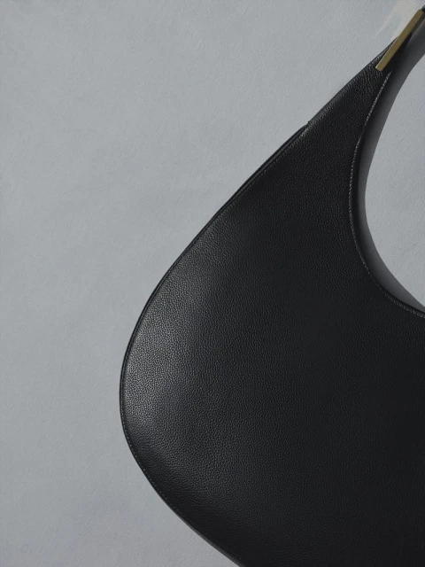 Close-up of a minimalist, black leather shoulder bag with a curved shape and a gold metal detail against a pale grey background.