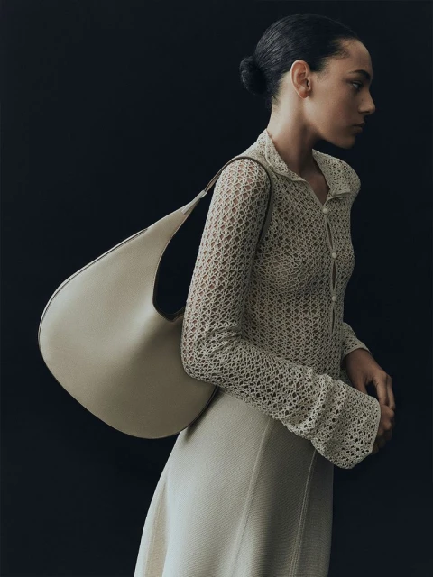 Woman poses with a beige hobo bag in a fashion studio shot. She wears a cream-colored knit shirt and skirt against a dark backdrop.