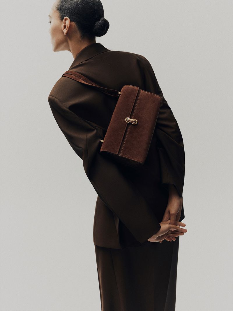 Model wearing an oversized dark brown blazer with a brown suede shoulder bag across her back against a studio backdrop.