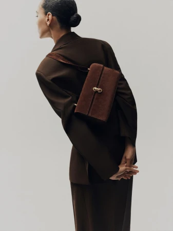 Model wearing an oversized dark brown blazer with a brown suede shoulder bag across her back against a studio backdrop.