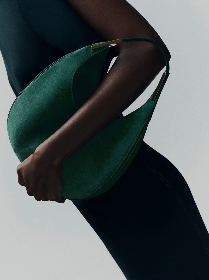 Dark-skinned model gracefully presents a green suede hobo bag with gold accents against a clean, light background in a fashion studio shot.
