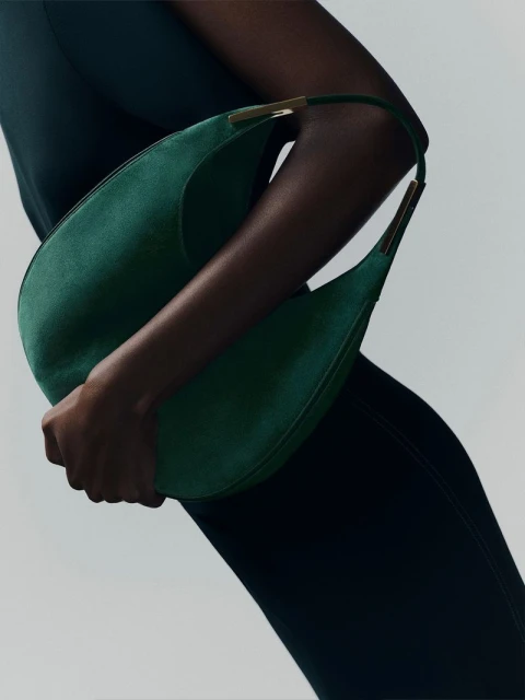 Dark-skinned model gracefully presents a green suede hobo bag with gold accents against a clean, light background in a fashion studio shot.