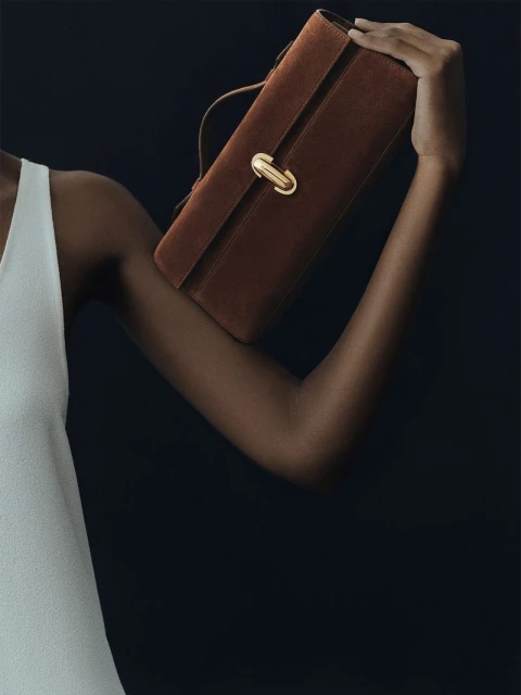Dark-skinned person in a white dress holds a brown suede clutch with a gold clasp against a dark background in a studio setting.