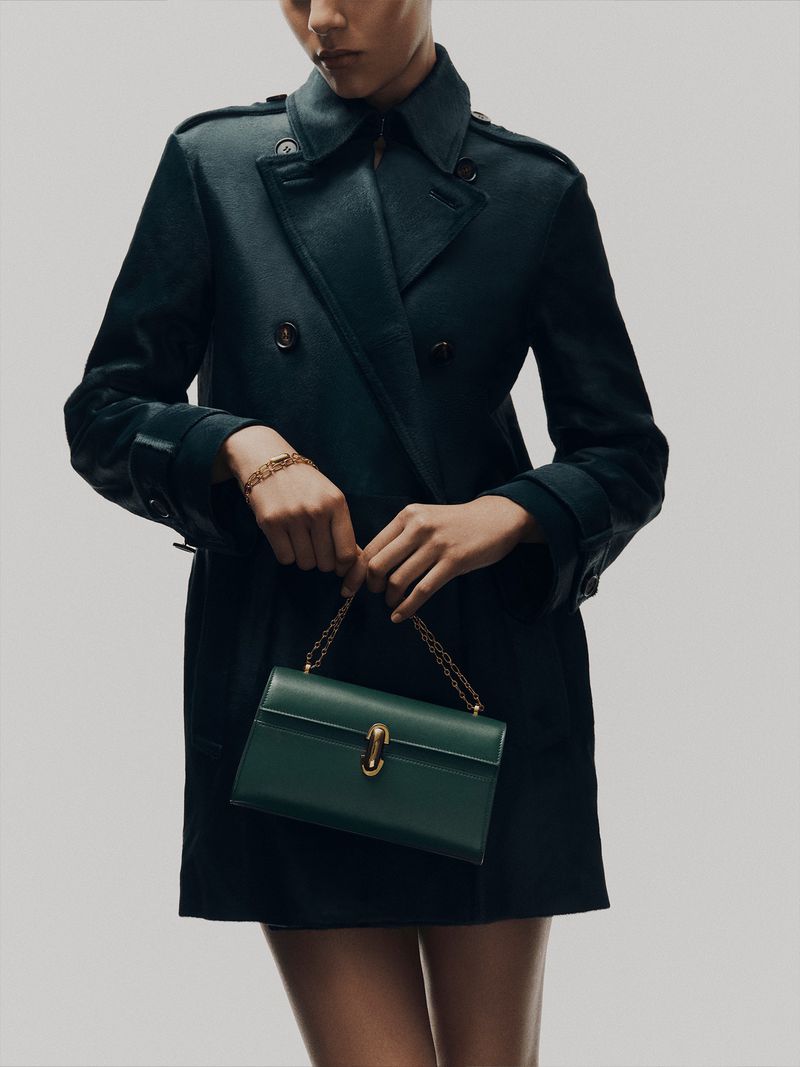 Model in dark green trench coat holding a matching green leather handbag against a light grey studio background.