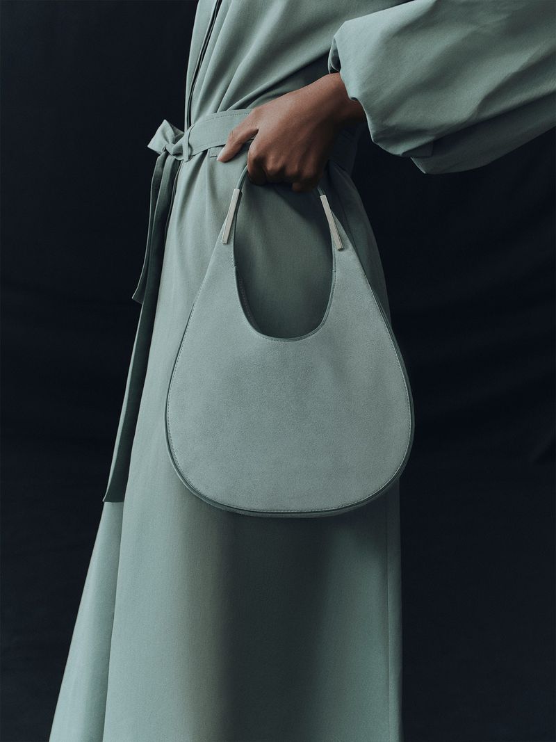 Woman in a pastel green trench coat holds a light green, rounded leather handbag against a dark gray backdrop in a fashion shot.