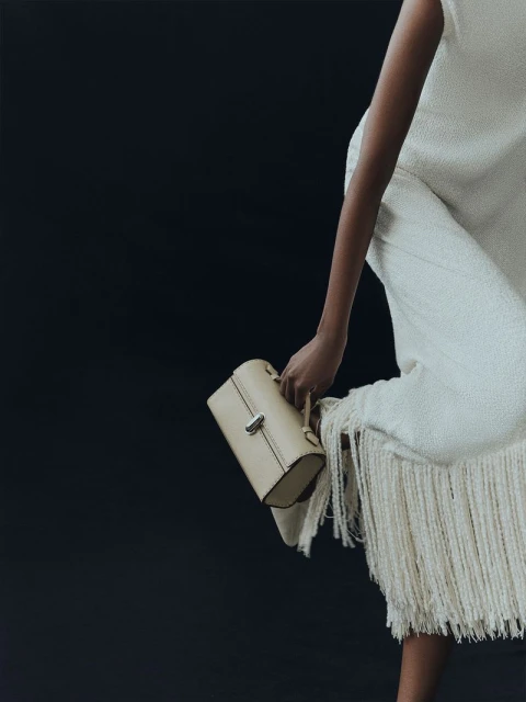 A woman showcases a handbag while sitting on a fringed garment against a black background in a studio fashion shoot.