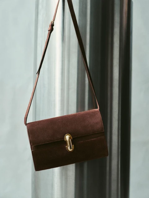 Dark brown leather purse with a gold clasp and a shoulder strap, displayed against a light grey background with a metallic column.
