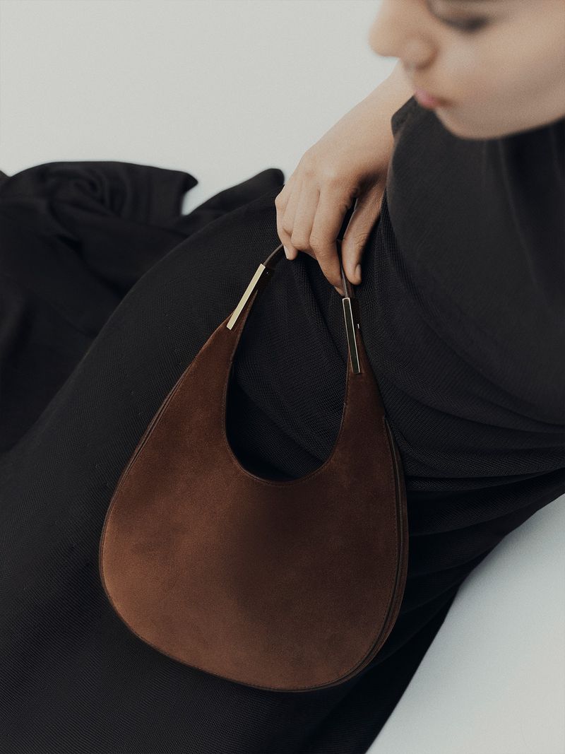 A brown teardrop-shaped suede purse with gold hardware is held by an adult female wearing a black dress against a white backdrop.