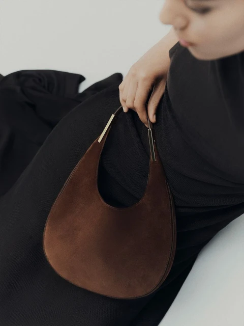 A brown teardrop-shaped suede purse with gold hardware is held by an adult female wearing a black dress against a white backdrop.