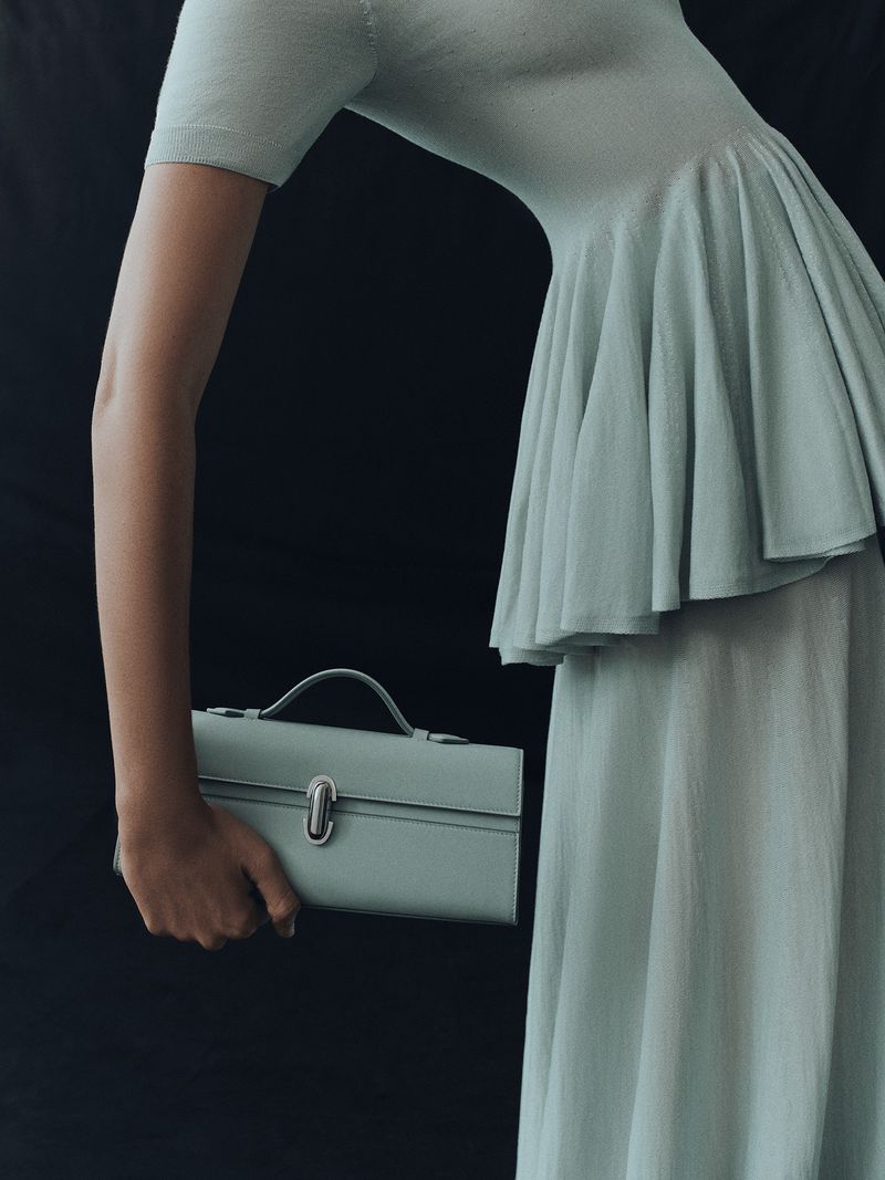 A woman in a light green dress holds a matching satchel against a dark background, showcasing fashion and style.