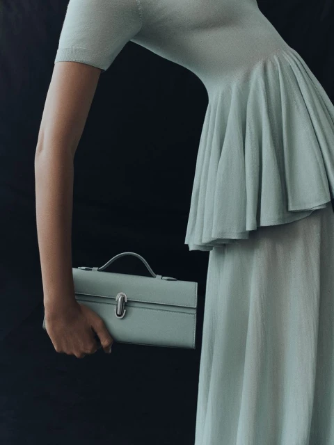 A woman in a light green dress holds a matching satchel against a dark background, showcasing fashion and style.