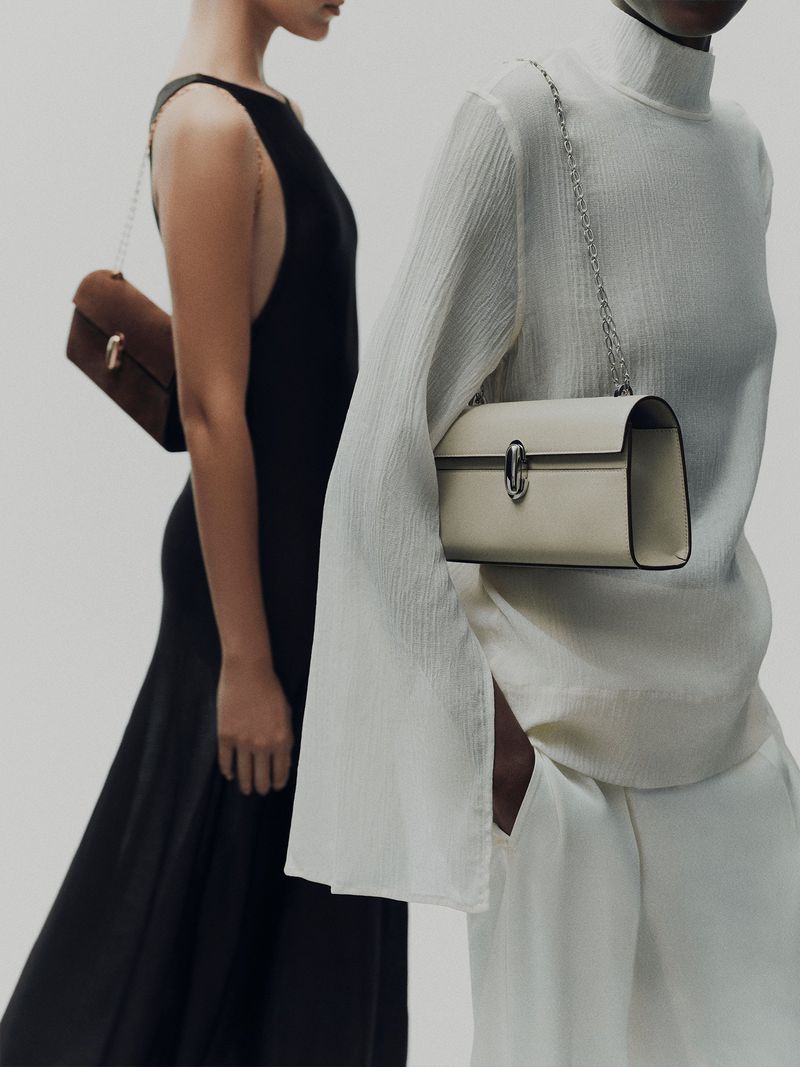 Two women stand in front of a white backdrop, each showcasing a stylish handbag with a chain strap. One woman wears a black dress.