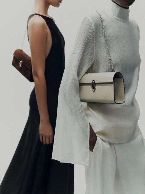 Two women stand in front of a white backdrop, each showcasing a stylish handbag with a chain strap. One woman wears a black dress.