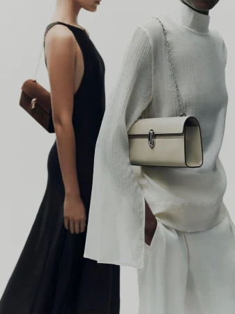 Two women stand in front of a white backdrop, each showcasing a stylish handbag with a chain strap. One woman wears a black dress.