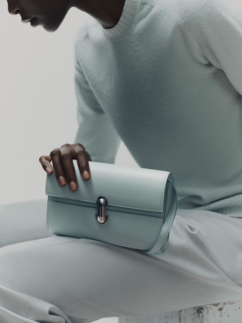 Close-up studio shot of a light-blue leather clutch purse held by a dark-skinned adult. The person wears matching light-blue knitwear.
