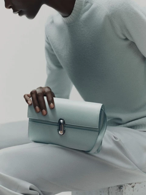 Close-up studio shot of a light-blue leather clutch purse held by a dark-skinned adult. The person wears matching light-blue knitwear.