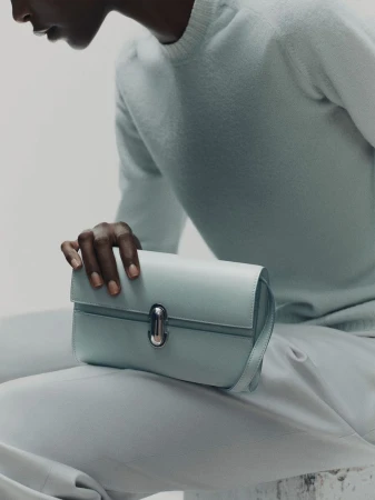 Close-up studio shot of a light-blue leather clutch purse held by a dark-skinned adult. The person wears matching light-blue knitwear.