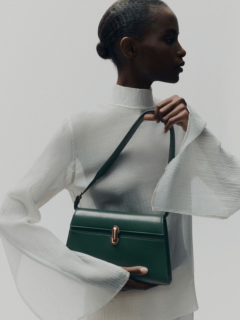 Elegant dark green leather satchel handbag displayed by a Black female model wearing a white top and neutral studio background.