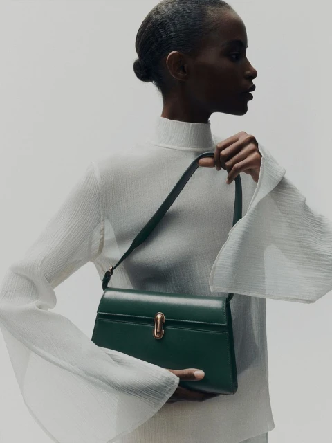 Elegant dark green leather satchel handbag displayed by a Black female model wearing a white top and neutral studio background.