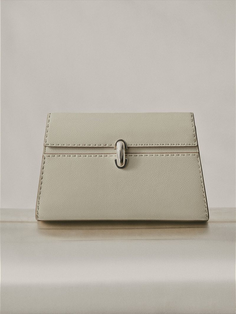 Elevated product shot of a pale grey clutch bag with a silver clasp on a light-colored surface in a minimalist indoor setting.