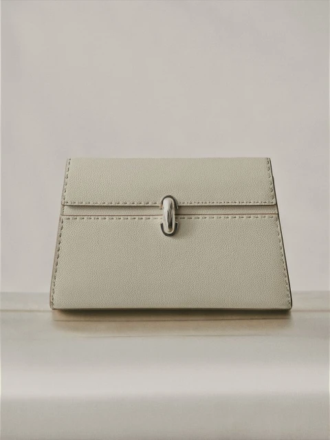 Elevated product shot of a pale grey clutch bag with a silver clasp on a light-colored surface in a minimalist indoor setting.