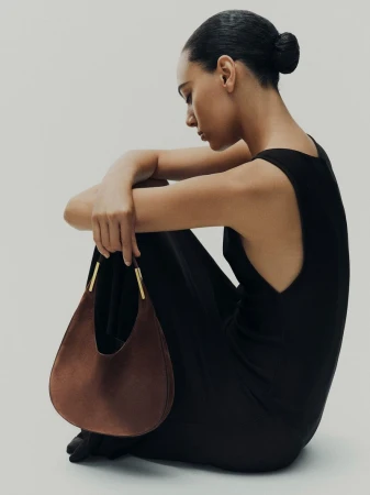 Model in a sleeveless black dress seated on the floor holding a brown suede crescent handbag with gold hardware.
