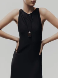 Cropped model in a black sleeveless dress wearing a brown leather pendant necklace against a light grey seamless backdrop.