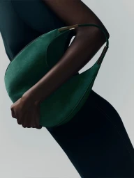 Cropped view of a model in a dark dress holding a structured emerald green suede shoulder bag with gold hardware against a grey backdrop.