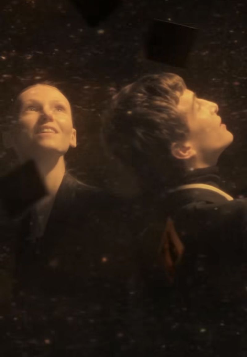 Two people, an adult male and adult female, look up in a dark environment illuminated with particles, wearing dark clothing, in a medium shot.