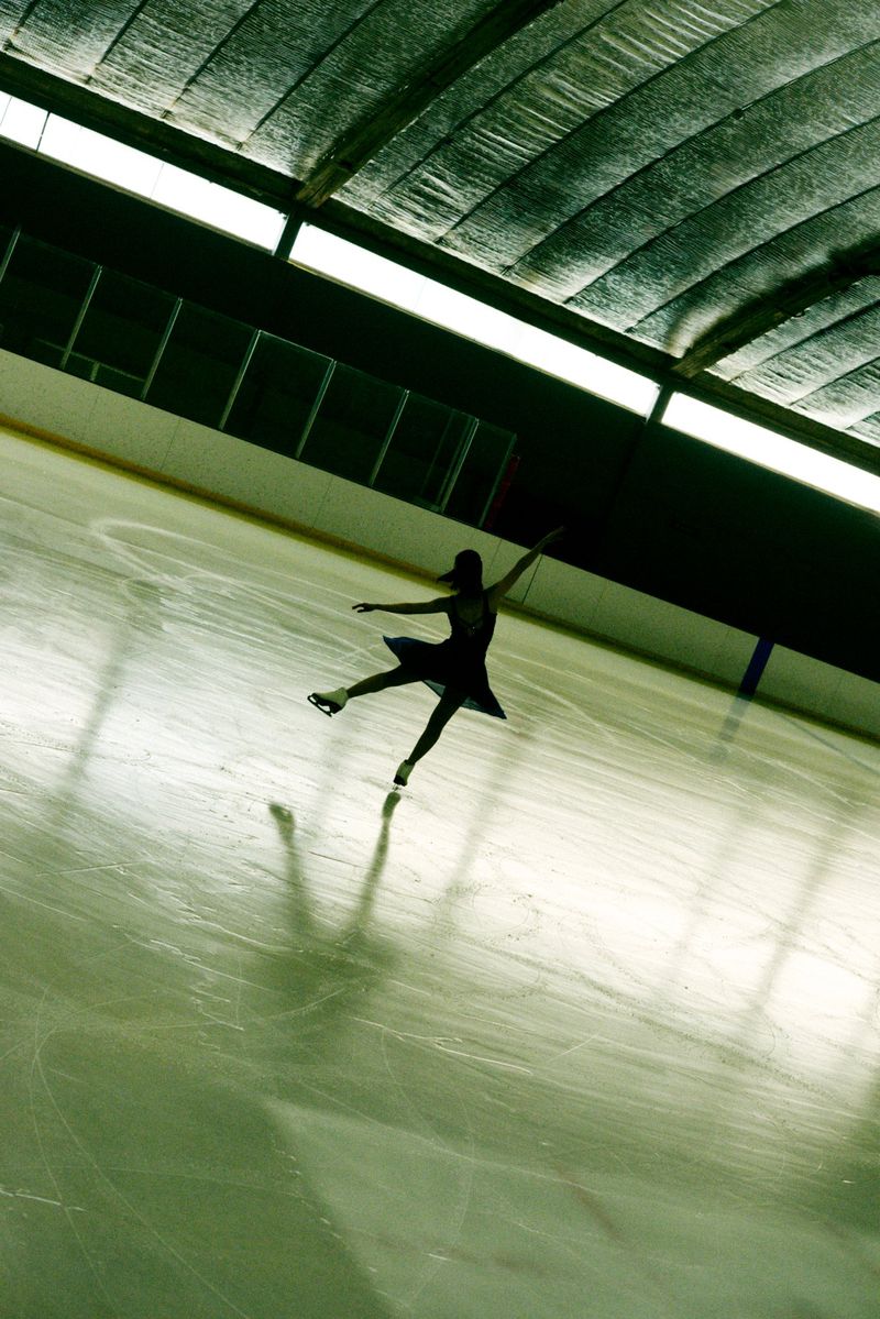 Solo female figure skater performing on an indoor ice rink with arms extended in a darkened arena with bright lighting.