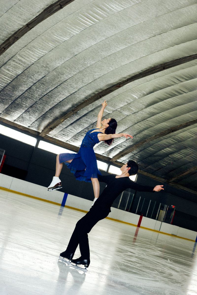 A pair of figure skaters, a man and a woman, perform a lift on an indoor ice rink, showcasing skill and athleticism under a high ceiling.