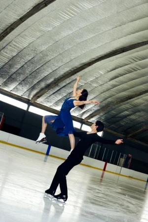 A pair of figure skaters, a man and a woman, perform a lift on an indoor ice rink, showcasing skill and athleticism under a high ceiling.