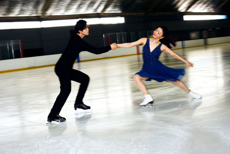 Figure skaters perform on an ice rink, holding hands and wearing costumes. The woman is in a blue dress, and the man is in black.
