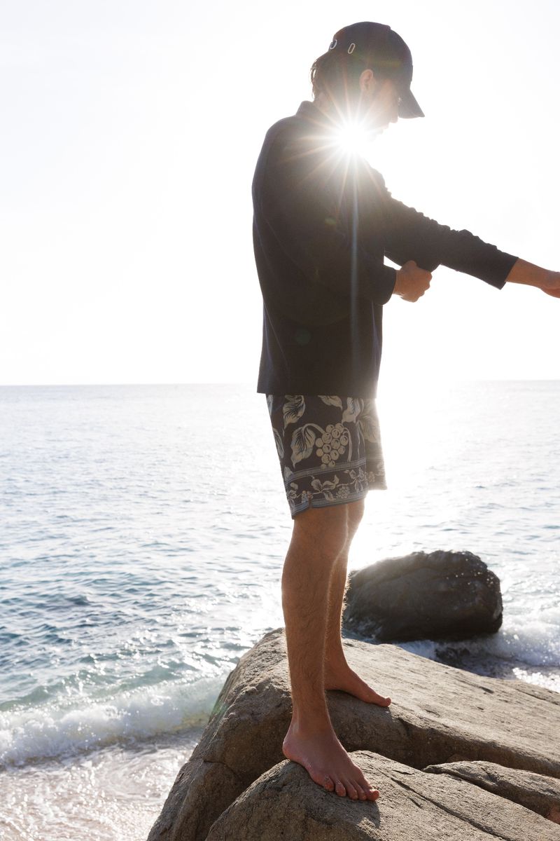 Person standing on rock by the sea, wearing a hat, dark jacket and floral shorts, backlit by the sun, with blue water and sky.