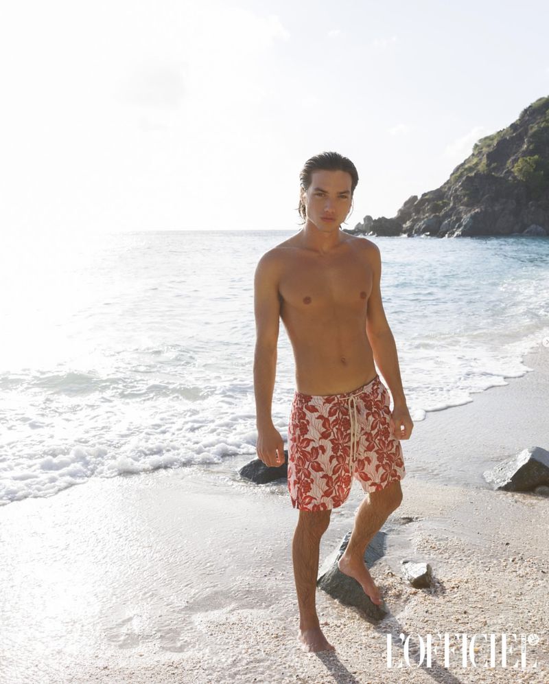 Male model poses shirtless on a sunny beach in floral swim trunks. Ocean waves break in the background, with lush green hills.
