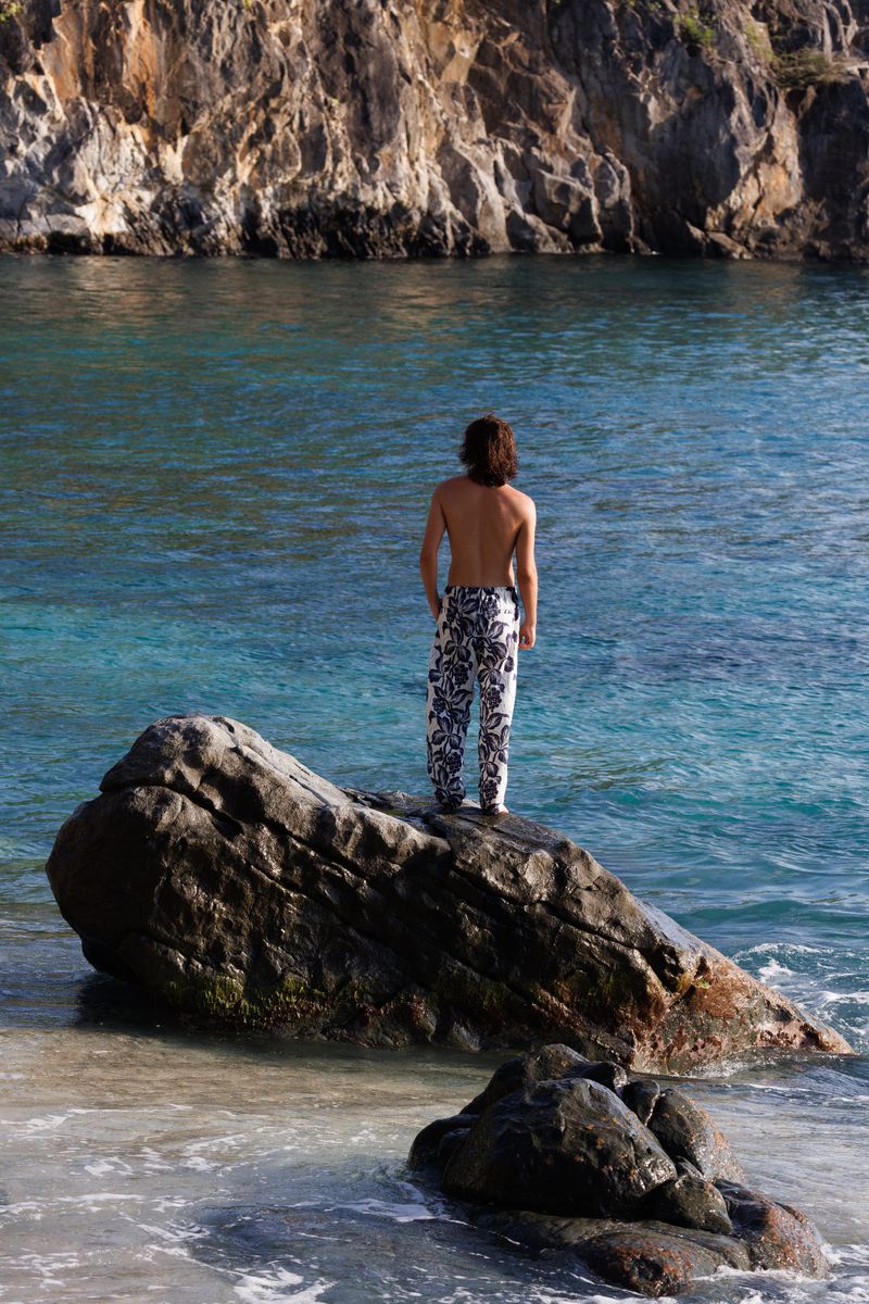 Shirtless man with long hair standing on a rock formation looking out at the ocean and rocky cliffs on a sunny day.