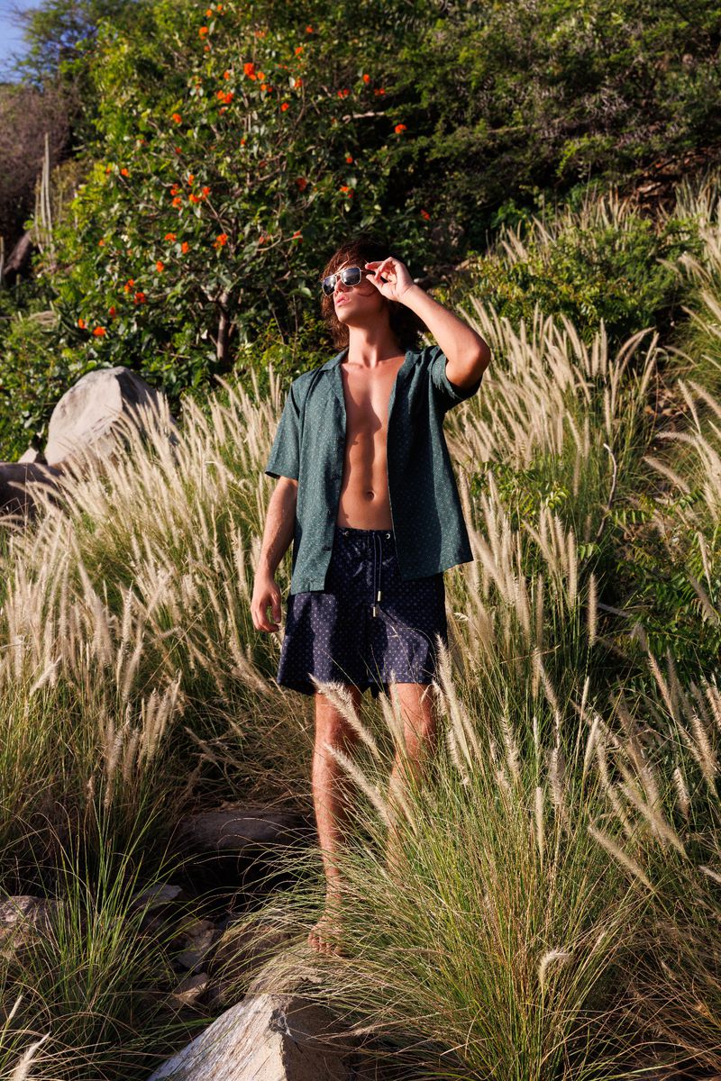 An adult male stands outside wearing an unbuttoned shirt, shorts, and sunglasses, surrounded by tall grasses and greenery.