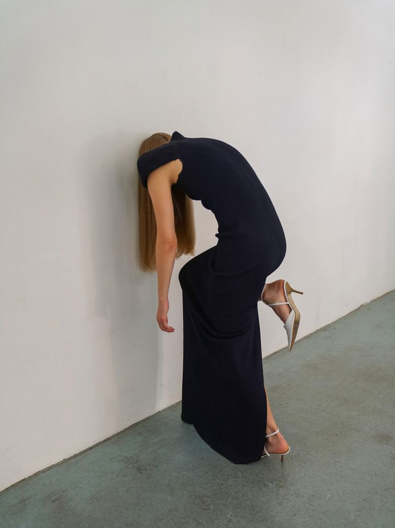 Adult female in a dark dress and light coloured high-heeled shoes, leaning against a white wall in an indoor environment. 