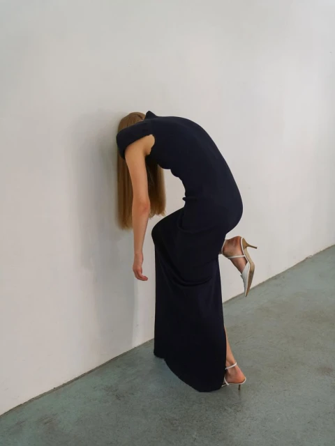 Adult female in a dark dress and light coloured high-heeled shoes, leaning against a white wall in an indoor environment. 