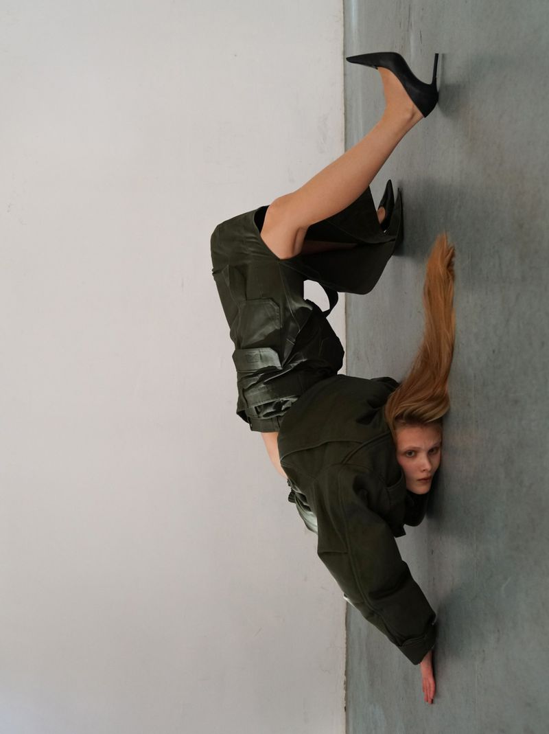 Blonde woman in green jumpsuit, black heels does handstand against grey wall. Fashion photography, indoor scene, hair flowing downward.