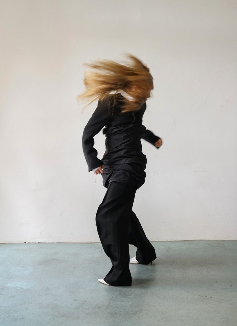Fashionable blonde-haired person in motion, wearing a black jacket, dark pants, and white pointed shoes against a light gray background.