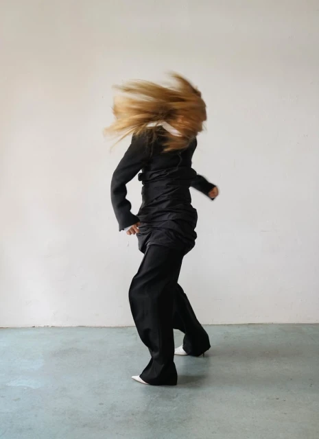 Fashionable blonde-haired person in motion, wearing a black jacket, dark pants, and white pointed shoes against a light gray background.